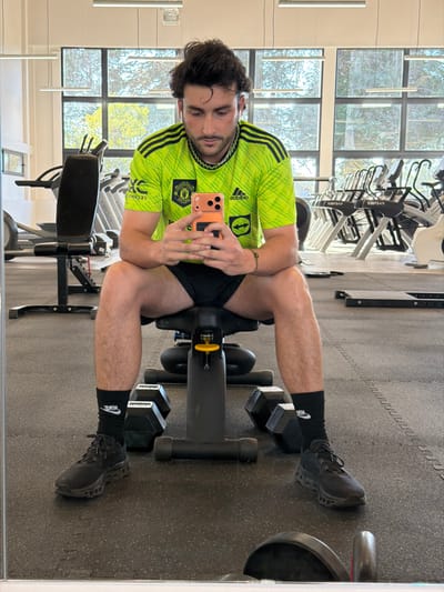 A man in a green and yellow shirt with black shorts sits on a leg extension machine, holding a phone up to take a selfie. He has dumbbells under each foot. He wears black socks and shoes. The image appears to be taken in a gym, reflecting in a mirror. Exercise bikes and other gym equipment are visible in the background. The lighting is bright, with natural light streaming through windows. The floor is covered in gray rubber gym flooring.
