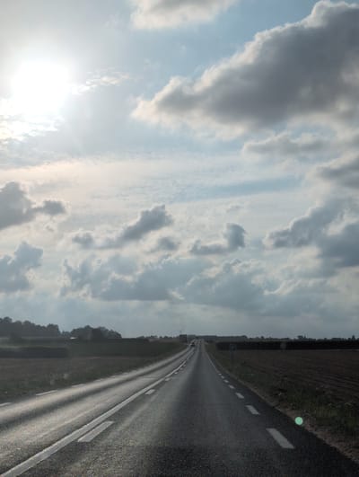 The image shows a paved road in Grigneuseville, France, under a cloudy sky with the sun partially visible and casting a bright glare. The road is lined with white markings and curves to the left in the distance. To the right of the road are tilled fields, and to the left, there are fields and trees. In the far distance, wind turbines are visible, and there is a dark silhouette of trees and foliage along the horizon. A car is faintly visible in the distance on the road. The overall scene depicts a rural landscape with a road leading through agricultural land.
