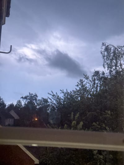 An outdoor scene captures a dusky sky with fragmented clouds, illuminated by soft twilight. A dark silhouette of dense trees forms the mid-ground, with the top branches of a weeping willow visible on the right. In the background, the faint outlines of houses suggest a residential area. A single, small, orange light source is visible amongst the trees. In the foreground, the edge of a building's roof and a section of brickwork are visible, indicating the viewpoint is from within a structure. The overall impression is of an evening scene in a suburban or semi-rural setting, likely within the Borough of Swale, United Kingdom.