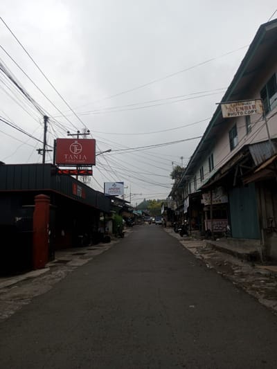 The image depicts a street scene in the city of Houaythat, Laos, under an overcast sky. A narrow, dark asphalt road extends into the distance, flanked by commercial buildings and utility infrastructure.
On the left side of the street, a dark-colored building with a prominent red pillar section is visible in the foreground. Above this building, a large red illuminated sign displays "TANIA PERFUME" with a stylized logo, below which is an LED ticker displaying red arrow patterns. A smaller sign below reads "WANGI ITU". A utility pole heavily laden with a tangle of black electrical and communication wires stands adjacent to this building. Further down the street on the left, an "APOTEK PRONESIA" (pharmacy) sign is visible, and several motorcycles are parked along the sidewalk. Indistinct human figures are present further along the street. In the distant background on the left, forested hills are visible.
On the right side of the street, a two-story building with a light-colored facade and multiple windows features a rusty metal sign hanging from its upper floor, which reads "TOKO BUKU DJEMBAR PHOTO.COPY" (Bookstore Djembar Photo.Copy). Below this, various shopfronts with corrugated metal awnings and shutters line the sidewalk. The concrete sidewalk on the right appears uneven and shows signs of debris and moisture. Motorcycles and possibly a car are parked further down the street on this side.
The sky is uniformly grey and overcast. Numerous overhead wires traverse the scene at various angles across the sky. The immediate foreground of the street and sidewalks shows some minor debris. No specific actions or interactions are clearly discernible among the distant figures.