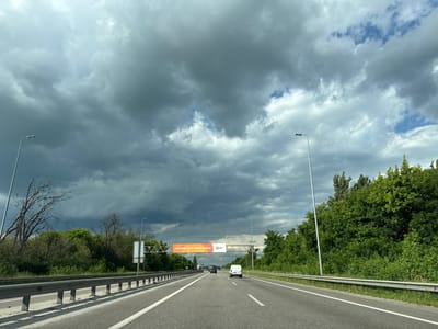The image depicts a view from a car driving on a highway in Ukraine. The scene is dominated by a dramatic, cloudy sky with dark storm clouds gathering in the distance, contrasted by patches of brighter sky. The highway itself is paved with white lane markings, and a metal guardrail runs along the side. Lush green trees and vegetation line the sides of the road, with a bare, dead tree visible on the left.

Several vehicles are on the road, including a white van in the right lane and a darker car further ahead. Overhead, a large banner is suspended across the highway. The banner features an orange and white design with text. The visible text on the banner reads "СИСТЕМНИЙ ЗАХИСТ ВАШОГО ВРОЖАЮ" and "UPL". The overall mood is somewhat somber due to the stormy weather, but the presence of the highway and vehicles suggests normal traffic flow. The time of day appears to be daytime, likely late afternoon given the lighting and the storm clouds. The location cues, such as the highway infrastructure and vegetation, are consistent with a rural or suburban setting along a major road in Ukraine.