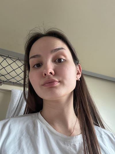 A young adult female is depicted in a close-up, head-and-shoulders shot, looking directly at the camera. She possesses long, straight, dark brown hair, dark brown eyes, and is wearing a plain white crew-neck t-shirt. Her right ear features a small hoop earring with a white spherical bead and a golden ear cuff on the upper cartilage. A thin, light-colored chain necklace is visible around her neck. The background shows the interior of a train cabin, characterized by a metallic mesh luggage rack on the left, below which a white towel is draped. A light beige ceiling occupies the upper portion of the background. The scene is located inside a train cabin, in the city of Barabinsk, Russia.
