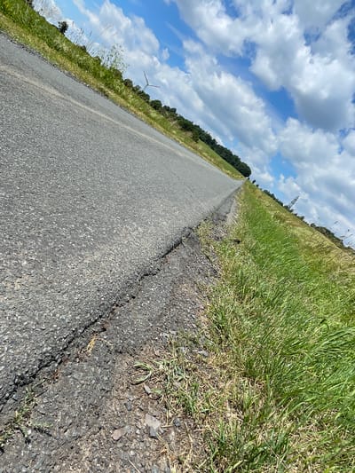 This is a low-angle, wide shot of an asphalt road in Schwarzenborn, Germany, angled diagonally from the lower left to the upper right. The road is cracked and appears to be in disrepair, with gravel and dirt along its edge. To the right of the road, tall green grass grows along a gentle slope. In the distance, a line of trees borders the field. The sky is a bright blue with scattered white, puffy clouds. A faint outline of a wind turbine is visible on the left side of the horizon, and a tall telecommunications tower is seen in the distance on the right. The scene appears to be in a rural or semi-rural setting.