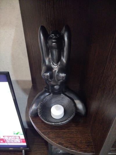 A dark, sculptural candle holder shaped like a woman in a kneeling pose with arms raised is displayed on a wooden shelf. The candle holder is made of a dark material and has a textured bowl in the center containing a white, unlit candle. To the left, a portion of a digital screen is visible, displaying a QR code and some text. The background consists of a dark wooden surface, likely part of a bookshelf or furniture. The image appears to be taken indoors, possibly in a home or a shop. There are no people visible in the image. The scene does not provide any specific information about the city of Nizhnyaya Maktauma, Russia.