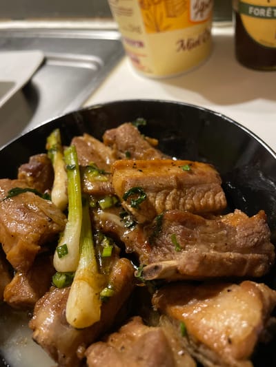 A close-up shot displays cooked pork ribs glistening with sauce, garnished with sliced scallions in a black bowl. The ribs exhibit a brown, caramelized appearance. In the background, partially visible, are two cylindrical containers. The composition suggests a focus on the savory dish, highlighting its visual appeal and fresh garnishing. The image provides a glimpse of the meal and associated condiment containers.