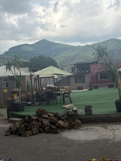 This image captures an outdoor scene in Goris, Armenia, with a backdrop of lush, rolling green hills and a cloudy sky. In the foreground, a pile of large, rough brown rocks sits on a gravel surface. To the right of the rocks, a paved area with a green artificial turf surface is visible, featuring a stacked pile of grey bricks and a concrete block bench.

Further into the scene, an outdoor seating area is arranged under a large, green umbrella. Several dark chairs surround tables, with one chair prominently placed to the left. A tall, slender palm-like plant in a dark pot stands to the left of the seating area. Behind the seating area, buildings with stone and red metal exteriors are visible. The buildings appear to be of a residential or commercial nature, with windows and some decorative elements.

The distant landscape is dominated by the verdant, tree-covered mountains characteristic of the Goris region. A prominent peak in the center distance features what appear to be communication towers. The overall impression is one of a semi-developed outdoor space with a natural, mountainous environment.