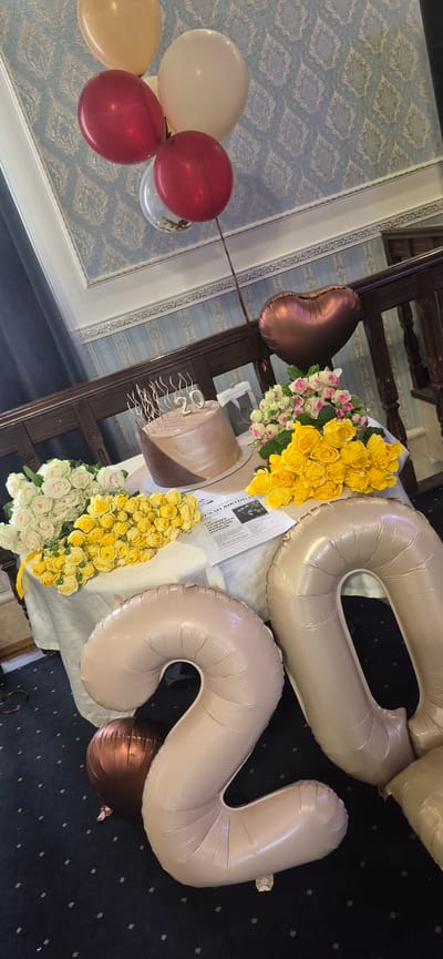 The image captures a festive scene, likely a birthday celebration, given the presence of balloons and a cake. Dominating the frame are two large, tan-colored foil balloons shaped like the numbers "2" and "0", suggesting a 20th birthday. Several smaller balloons in cream, red, and possibly gold are clustered together on the left side of the image.
A two-tiered cake, adorned with what appears to be chocolate frosting, sits on a table. Delicate golden numerals, also reading "20", are placed on top of the cake as a decorative element. Flanking the cake are generous bouquets of yellow and white roses, with a smaller cluster of pink and white roses to the left. A heart-shaped rose gold balloon is positioned near the smaller rose bouquet.
The background features a patterned wallpaper in pale blue and white, and dark wooden furniture, possibly part of a dining room or a hall. A dark carpet is visible in the lower right portion of the image. A newspaper clipping, with text visible, is placed under the cake and balloons. One headline that is partially legible reads "IT'S MY BIRTHDAY". The overall impression is one of a carefully arranged and joyous celebration. The lighting suggests it is daytime. No people are visible in the image.