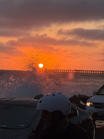 A dynamic sunset over the Pacific Ocean in Oceanside, California is captured in this vertical, medium-shot image. The sun, a bright, incandescent orb, is positioned near the center of the frame, low on the horizon. It is surrounded by a dramatic explosion of water droplets, creating a spray that catches the sunlight and glistens. The sky above is a gradient of deep purple at the top, transitioning to fiery oranges and pinks as it nears the horizon, with scattered clouds adding texture.

In the foreground, the silhouettes of several parked vehicles are visible. One car, a dark sedan, occupies the lower left and center of the frame, its windshield reflecting the twilight sky. A white SUV is parked to the right, its headlights illuminated. In the immediate foreground, a person wearing a white helmet is partially visible, facing away from the camera and towards the ocean.

A long pier, lined with lampposts and dotted with silhouetted figures, stretches across the middle ground of the image, extending into the ocean. The water itself is a deep, reflective blue, with small waves breaking and adding to the overall sense of movement and energy in the scene. The shoreline appears to be rocky, with dark boulders visible in the foreground. The overall impression is one of a powerful and vibrant natural display at dusk, with human activity subtly present.
