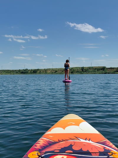 A person stands on a pink paddleboard in the middle of a calm body of water. The person is wearing a dark blue swimsuit and is holding a paddle. The water has small ripples and reflects the blue sky with scattered white clouds. In the background, a tree-lined shore with rolling hills is visible. Electrical poles are scattered along the top of the hills. The foreground shows the front of another paddleboard with an orange and red design featuring a stylized face. The image appears to have been taken on a sunny day in Samoilov, Russia, likely during recreational activity.