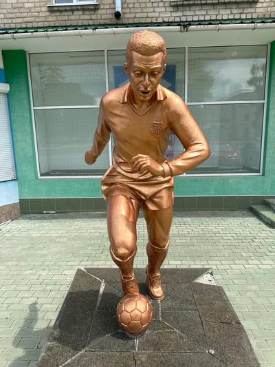 This is a full-body, low-angle shot of a bronze statue of a soccer player in action, positioned on a dark granite pedestal. The statue depicts a man with short, textured hair, a determined expression, and muscular build, wearing a short-sleeved jersey with a crest on the chest, shorts, and cleats. His left leg is bent and positioned behind him, while his right leg is extended forward, kicking a soccer ball at his feet. The statue is golden in color, suggesting bronze.

The setting appears to be outdoors, in front of a building with large windows and green-painted walls. The ground is paved with light green, diamond-shaped tiles, with a darker paved area around the pedestal. In the background, reflections of trees and sky are visible in the windows. A white pipe runs vertically along the brick wall to the left of the statue.

The activity depicted is a soccer player in the process of kicking a ball. The time of day seems to be daytime, with sunlight casting shadows on the ground. The weather appears to be clear, as indicated by the bright light and reflections in the windows. The location is identified as Staryi Aidar, Ukraine. The crest on the statue's jersey is partially visible, but too indistinct to read.