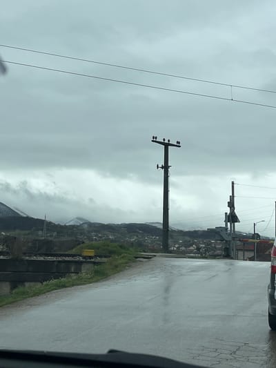 The image shows a scene in or near Cacak, Serbia, on a cloudy day, seemingly captured from inside a vehicle moving along a wet, cracked road. A utility pole with wires is visible to the left of the road. On the right side of the road, a grassy verge transitions into a hillside populated with buildings and vegetation. In the distance, mountains are visible, obscured by heavy cloud cover. A railway crossing barrier is seen up ahead, and the camera car is likely at a railway crossing in the region.