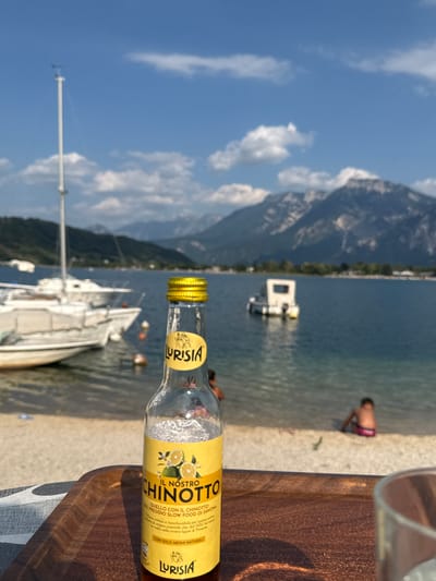 The media file captures a scenic view of a lake with mountains in the background, under a clear blue sky with scattered clouds. A beach with a few people is visible along the shoreline. In the foreground, a glass bottle of "IL NOSTRO CHINOTTO" by Lurisia is prominently displayed. The bottle has a yellow label featuring illustrations of chinotto fruits and text in Italian. A person wearing a red and black swimsuit is sitting by the water's edge, looking away from the camera. Further out on the lake, several sailboats are moored, and a small white structure resembling a floating platform or buoy is visible. The overall impression is a relaxed, summery day in a picturesque lakeside location, likely in the late afternoon given the lighting. The visible text on the bottle translates to "Our Chinotto, the one from the Slow Food Presidium of Savona. A unique and unmistakable citrus fruit from the Ligurian Riviera. Its intense and unforgettable flavor, dating back to the 16th century, comes from the rare oriental chinotto fruit, grown in the western Ligurian Riviera."