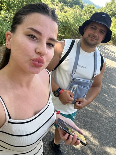 A young woman in a white and black striped tank top is taking a selfie outdoors, puckering her lips towards the camera. She has dark hair pulled back and is wearing a small silver earring. Beside her, a man in a white sleeveless shirt and a black bucket hat smiles at the camera. He is wearing a grey crossbody bag and a red and black watch. The background shows a wooded hillside with green foliage and a paved path. The setting appears to be a natural environment, potentially a park or hiking trail. No specific landmarks within the city of Kremen, Bulgaria, are discernible in the image.