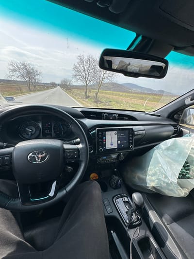 The image depicts a first-person perspective from the driver's seat of a Toyota vehicle, likely stationary or moving slowly, on a rural road in the vicinity of Zlati Voyvoda, Bulgaria. The driver's legs, clad in dark trousers, are visible in the lower left. The black steering wheel, centered in the frame, features the Toyota emblem. The vehicle's instrument cluster indicates a digital odometer reading of 18,949 km. To the right of the steering wheel, the infotainment system displays a navigation map showing a route with "Sofia, Bulgaria" as a destination, 48 km away. The automatic gear selector is positioned in "P" (Park).
A large, translucent light-blue plastic bag containing green, foliage-like material, possibly artificial plants or decorative items, occupies the front passenger seat. A dark accessory hangs from the center console, and a pair of silver-framed eyeglasses rests in a storage compartment within the center console. The rearview mirror reflects elements of the vehicle's interior.
Outside the vehicle, a straight, two-lane asphalt road recedes into the distance under a bright, partly cloudy sky. The roadside features dry grass and sparse green vegetation, flanked by numerous leafless deciduous trees, suggesting late winter or early spring. Beyond the immediate roadside, cultivated fields with distinct patches of brown and green stretch towards a range of distant hills or low mountains on the horizon. The landscape is predominantly rural and open.