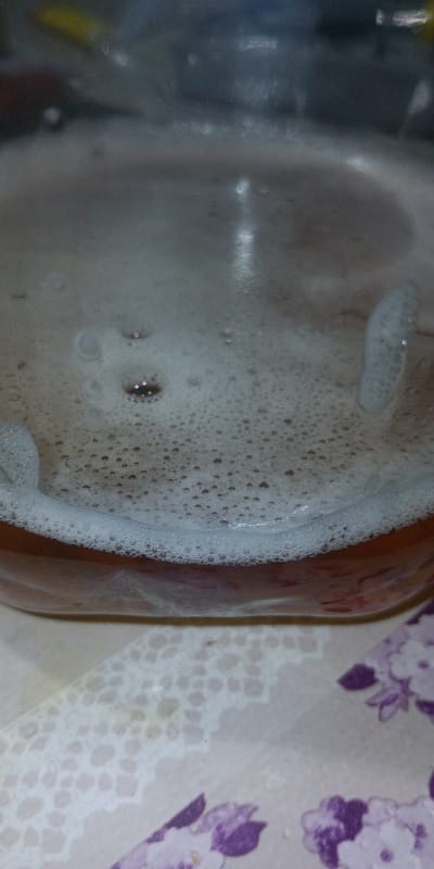 The image shows a close-up view of a clear glass jar containing a foamy, brown liquid. The liquid has a layer of small bubbles on its surface, with larger bubbles concentrated near the rim of the jar. The jar is placed on a patterned surface, possibly a tablecloth, with a floral design featuring purple and white elements. The background is dark and out of focus, with some hints of yellow and blue. There are no people visible in the image. This scene is not located in Gornyak, Russia.