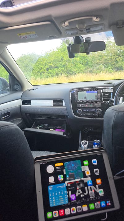 The image shows the interior of a car, viewed from the back seat, with a tablet displaying the home screen positioned in the foreground. The car appears to be parked on a roadside with a grassy verge and trees visible through the windshield. The tablet displays various app icons and widgets, including a clock, weather forecast, and a map. In the center console of the car, a gear shift, a can of soda, and a charging port are visible. The glove compartment is open, revealing some items inside. The overall scene suggests the car is stationary, possibly during a break or a stop. The reference to Wicken, United Kingdom, is not directly ascertainable from the visual information provided in the image.