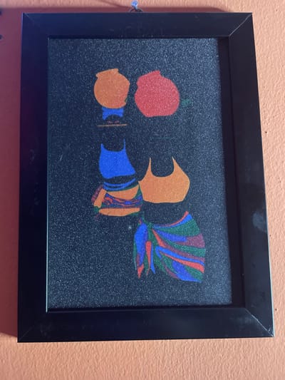 A framed print with a black background features abstract shapes in vibrant colors. A red shape resembles a fruit or flower. An orange figure has beak-like edges, positioned next to blue shapes that could represent a body or garment. Rainbow-colored lines flow from one edge, adding movement and interest to the composition. The image is clean, minimalist, and visually striking.