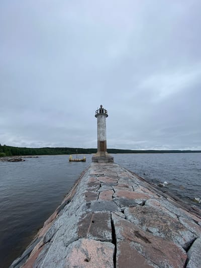 A lighthouse stands on a stone pier extending into dark, choppy water under an overcast sky. The lighthouse is cylindrical and made of concrete with a metallic top. A wooden door is visible at the base of the lighthouse. The pier is constructed from large, rough-hewn stones, some of which are covered in lichen. To the left of the pier, a small, ornate boat with figures and decorative elements is moored. The background features a dense forest lining a sandy shoreline, with more rocky outcrops visible on the left. The overall scene conveys a sense of remoteness and an industrial maritime setting. The location is the lighthouse on the pier in Motornoe, Russia.