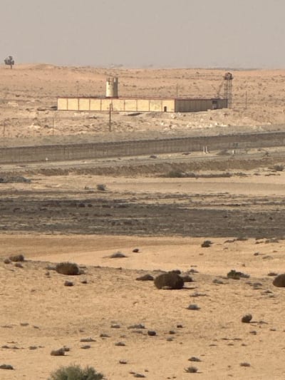 The image displays an arid desert landscape within the Ramat Negev Regional Council, Israel, under a hazy, pale sky. In the background, a long, rectangular building with light-colored walls and a darker roof is prominently situated on a raised, rocky area. A circular watchtower with crenellated top sections stands at the building's left end, while a taller, metal-framed observation or utility tower is visible on the right. A single, small tree is discernible on a distant hill to the far left. The midground features a multi-layered security barrier, likely a fence with a concrete base, spanning horizontally across the terrain, running parallel to an unpaved track or road. The foreground consists of sandy, rocky ground sparsely covered with low-lying scrub vegetation. No individuals, specific actions, or interactions are visible within the frame.