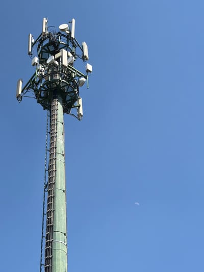 A low-angle shot shows a communication tower set against a clear blue sky, taken in Gudo Visconti, Italy. The top of the green, cylindrical tower is densely packed with multiple antennas and electronic equipment. A slender crescent moon is visible in the upper right portion of the frame. The tower is a mesh of cables and supportive framework. The background consists only of the sky.