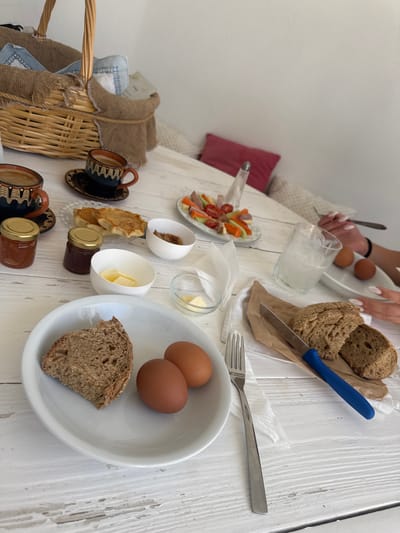 An overhead shot captures a rustic breakfast spread laid out on a weathered white wooden table, likely in Naousa, Greece. The scene is bathed in natural light, suggesting an outdoor or well-lit indoor setting. In the foreground, a white plate holds a slice of dark bread and two brown eggs, accompanied by a fork. To the right, a brown paper wrapper displays more dark bread, a knife with a blue handle, and a white napkin. A glass of light-colored liquid sits nearby, and further back, a small dish of butter is visible. Two small jars, possibly containing jam, are placed beside patterned coffee cups and saucers, hinting at a traditional Greek breakfast. A wicker basket, lined with burlap and containing folded napkins, rests in the upper left corner. In the background, a platter of colorful sliced vegetables and meats, along with a salt shaker, suggests a shared meal. The presence of hands, one holding a fork and another a knife, indicates that the meal is either in progress or about to begin.