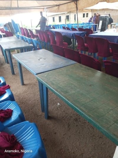 Under a tent, rows of blue and maroon chairs stand ready for an event. The chairs are not occupied, some adorned with maroon fabric. In front of them, tables line up, some covered in blue cloths. A few individuals are visible in the background, adding a sense of anticipation to the scene. The setting, likely for a gathering, is marked by a combination of rustic and festive elements. The image is tagged "Aramoko, Nigeria".
