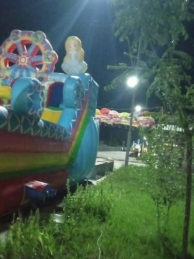 The image captures an outdoor scene at night, likely in the city of Dustlik, Uzbekistan, showcasing elements of an amusement or event area. In the foreground, a large, inflatable structure with vibrant colors is prominently featured. This structure appears to be part of a bouncy castle or similar attraction, with inflatable pillars and a colorful Ferris wheel design. A doll-like figure with blonde hair is situated atop a section of the inflatable. To the right, a tree with dense foliage is visible, partially obscuring a lamppost with a bright light. In the background, a row of colorful umbrellas are suspended, suggesting a festive or market atmosphere. The ground is covered with grass and some scattered debris. The overall impression is one of a lively nighttime gathering or entertainment venue.