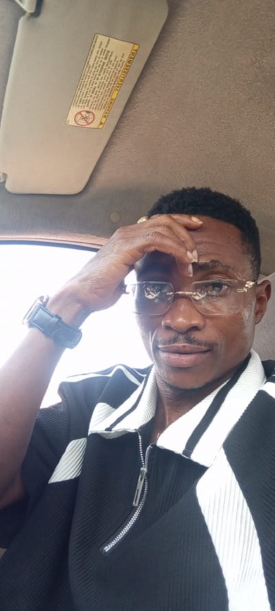 An adult male is depicted in a medium close-up photograph taken inside a vehicle in Osara Gada, Nigeria. The subject, with dark, short, curly hair and a mustache, wears rimless eyeglasses with gold-colored hardware. He is dressed in a black, ribbed, short-sleeved polo shirt featuring a white ribbed collar, white vertical panels, and a silver-colored quarter-zip closure. A black watch with a square face is present on his left wrist. His right hand is positioned with fingers splayed, resting on his forehead, and a gold-colored ring is visible on his right ring finger. He maintains direct eye contact with the camera, exhibiting a slight smile. The vehicle's interior features a beige/tan headliner and an upright sun visor on the passenger side, which bears an inverted yellow warning sticker with black text and an airbag safety symbol. A bright, diffused light source, indicative of an outdoor daytime setting, is visible in the upper left of the frame.