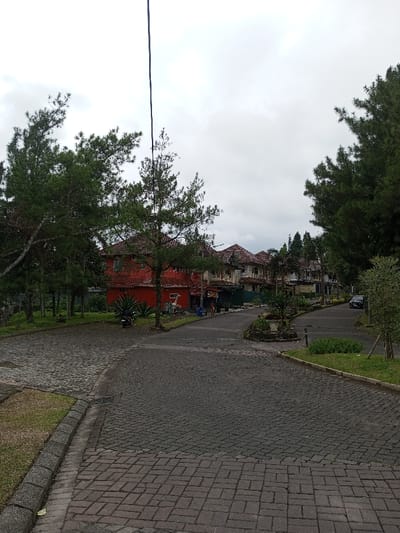 A quiet neighborhood street scene features houses with red-tiled roofs nestled amongst tall pine trees under an overcast sky. The roadway is paved with cobblestones. A motorcycle is parked near a red building. A car is visible further down the road. The overall setting suggests a tranquil residential area, possibly in a hilly or mountainous region.