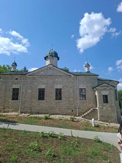 The image depicts a stone building with a green domed roof and two smaller green domed structures topped with crosses, suggestive of a church or monastery. The building's facade is constructed from rough-hewn light-colored stones, with several windows. A set of stone stairs leads up to an entrance on the right side of the building. A paved walkway is visible in the foreground, bordered by grassy areas and soil with some small plants. In the background, trees are visible. The sky is blue with scattered white clouds. The scene appears to be outdoors on a sunny day. The building is identified as the Condrița Monastery in Condrița, Moldova. A portion of a person with dark hair is visible on the far right edge of the image.