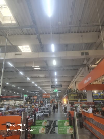 The image depicts the interior of a large retail store, likely a hardware or DIY store, with high ceilings and fluorescent lighting. Various aisles of merchandise are visible, including shelves stocked with products. Overhead signs indicate departments such as "Farben / Tapeten" (Paints / Wallpaper), "Auto / Fahrrad" (Car / Bicycle), and "Information." Several people are present, some browsing merchandise and others at a checkout counter. A bicycle is displayed as a product or promotion. The store appears to be well-lit and organized. The timestamp "12. Juni 2026 12:47" is overlaid on the bottom left corner of the image, along with "Galaxy S20 FE". The store is located in Gensingen, Germany.