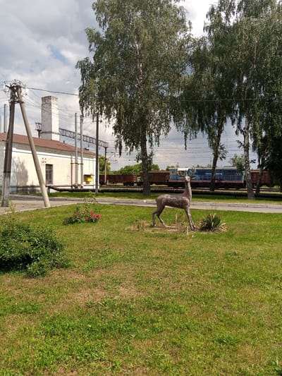 In Romodan, Ukraine, a statue of a deer stands on a grassy area in front of a building and railway tracks. The building, likely industrial, has a light-colored facade and a red roof, with industrial chimneys visible. Power lines and poles are present near the building. Behind the building, a train with several cars, including a blue locomotive, is parked on the tracks. The deer statue is positioned to the right of the frame, facing left, with its antlers visible. Green grass covers the foreground, with a small cluster of pink flowers near the left edge of the statue. The sky is partly cloudy.