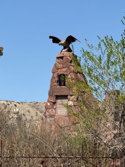 The image, correctly oriented, displays an outdoor scene in Barbulak, Kyrgyzstan, featuring a monument under a bright, clear blue sky. The primary subject is a large, bronze or dark-colored statue of an eagle with outstretched wings, appearing to land or take flight. This eagle statue is prominently mounted on a robust monument constructed from reddish-brown, rough-hewn stones.
To the right of the eagle statue, embedded within the stone monument, there is a rectangular plaque or recess with a dark surface. Further right on the stone pillar, an inscription in Cyrillic script is carved. The most legible text reads "СПАСИБО ЗА ПАМЯТЬ" (Thank you for the memory). Below this phrase, additional lines of Cyrillic text are visible but less distinct, including what appears to be the year "1941" and fragmented words such as "...ВТЕРИ".
The monument is partially framed by nature. From the upper left, branches with vibrant green leaves from a tree extend into the blue sky. On the right side of the image, below the main monument, a lighter-colored stone wall or structure is visible. The foreground on the right is dominated by a dense growth of dry, twiggy vegetation, intertwined with a rusty, metal fence-like structure or rebar.
The clear blue sky and strong illumination indicate a sunny daytime setting. The monument, with its eagle statue and inscriptions, clearly serves as a memorial, likely commemorating an event related to the year 1941, possibly World War II. The eagle symbolizes strength and national identity, common motifs for such sites. No human figures or activities are depicted.