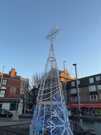 The image depicts a large, conical, artificial Christmas tree structure illuminated with white and blue LED lights and snowflake motifs, topped by a prominent snowflake ornament. The scene is set in Weymouth, United Kingdom, at an urban intersection. To the left, a multi-story brick building houses "The Somerset House Hotel," identifiable by signage. To the right, a building with a darker facade contains "Park Corner Convenience Store" at ground level, also indicated by signage. Behind the central tree structure, a taller building with scaffolding on its upper levels and roof is visible. A clear blue sky is overhead, suggesting daylight. One individual is visible walking near the convenience store on the right. A dark car is parked on the street adjacent to the hotel, and a white van is partially visible further down the street on the right. Black metal railings are in the immediate foreground. Deciduous trees in the background are bare, indicating a late autumn or winter season.