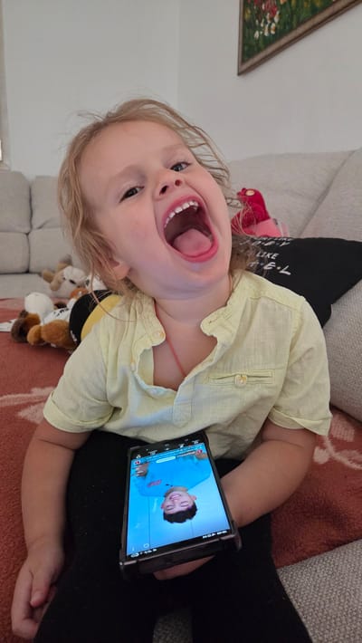 The image features a young child with blonde hair, wearing a light yellow short-sleeved shirt, lying down. The child's mouth is wide open in a laugh or shout, and their eyes are looking upwards. The child is holding a smartphone in their right hand, which displays a video of a young person with dark hair wearing a blue t-shirt. The background shows a couch and a textured blanket, suggesting an indoor setting. Several plush toys are visible near the child's torso, including one that appears to be a bee. A framed picture with greenery and flowers can be seen in the upper left corner. The lighting suggests it might be daytime. The overall emotion conveyed is one of joy and engagement. The image is oriented sideways.