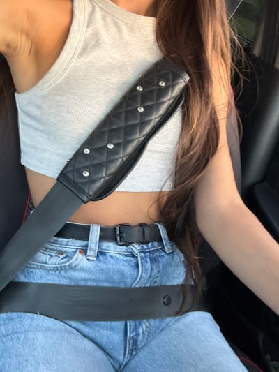 The image shows a close-up of a person sitting in the driver's seat of a car. They are wearing a light grey crop top, blue denim jeans, and a black belt. A black seatbelt is across their chest, with a quilted black padded cover adorned with small rhinestones. The person has long, dark brown hair. The interior of the car, including a black car seat and part of the steering wheel, is visible. The lighting suggests it is daytime. There are no discernible landmarks or clues within the image to indicate a specific location in Sadaclia, Moldova.