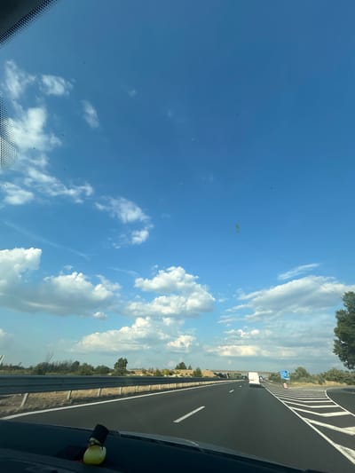 The image depicts a view from inside a vehicle traveling on a highway. The foreground shows a portion of the dashboard with a yellow rubber duck wearing a black top hat and sunglasses. The highway stretches ahead, with multiple lanes marked by white lines. A metal guardrail runs along the side of the road, separating it from the surrounding landscape. The landscape consists of sparse vegetation, trees, and a bridge or overpass in the distance. The sky is bright blue with scattered white cumulus clouds. A white van is visible on the highway ahead, and several road signs are present. The scene is bathed in daylight, suggesting it is daytime. There are no people visible outside the vehicle. The specific location in Tsalapitsa, Bulgaria is not ascertainable from the image itself.