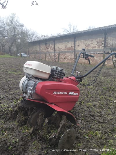 The image shows a red and white Honda FG320 tiller resting in muddy soil. The tiller is positioned in an outdoor area with a brick and wood fence in the background. Bare trees are visible in the distance, and a car can be seen behind the fence. The location is identified as Balgarsko Slivovo, Bulgaria, and the date and time are indicated as March 29, 2026, at 09:31.