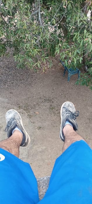 A person wearing blue shorts and gray shoes stands on a dirt path. Green foliage lines the left side of the path and a blue, overturned chair is slightly obscured in the green area. The path is composed of packed dirt and small pebbles. The photograph is taken from a high angle, looking down at the person's legs and the ground.