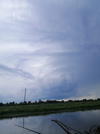 The image depicts a calm body of water reflecting a cloudy sky and a treeline in the distance. In the foreground, a fishing rod is partially submerged in the water, with another fishing rod visible to the right. A tall wooden pole stands on the grassy bank beyond the water. The sky is filled with varying shades of grey and blue clouds, suggesting an overcast or approaching storm. The environment appears to be a natural or rural setting. The location is identified as Krasnoluka, Ukraine. There are no people visible in the image.