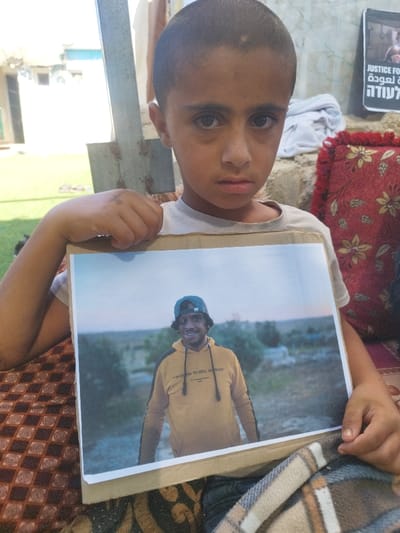 A young boy is depicted seated outdoors in Carmel, Palestinian Territory, holding a framed color photograph. The boy, who appears to be of elementary school age, has short dark hair and dark eyes, and is wearing a light-colored short-sleeved shirt. He looks directly at the viewer with a serious expression.

The photograph he holds, approximately A4 size and mounted on a cardboard backing, displays a smiling young man. This man wears a yellow hooded sweatshirt with black accents and the partially visible English text "I'M GOING TO HELL ANYWAYS" printed on the front. He also wears a dark blue baseball cap, reversed, and has dark hair and a beard. The background within the photograph features an open, semi-arid landscape with sparse vegetation and a dirt path under a pale sky, suggesting dusk or dawn.

The boy is seated on a patterned reddish-brown mat or rug, covered in part by a plaid blanket with brown, tan, and blue checks. The immediate background to the boy's right (viewer's left) includes a bright, sunlit area, possibly an opening, and a vertical metal beam. Behind the boy, a light-colored block or stone structure is visible, along with draped white fabric. To the boy's right (viewer's right), a decorative red cushion with gold floral patterns rests against the background. Further back, a black poster with white text is affixed to a surface. The poster displays English text reading "JUSTICE FOR" and "THE RETURN," along with prominent Arabic script, including "العودة" (Al-Awda, "The Return"). The scene is illuminated by natural daylight.