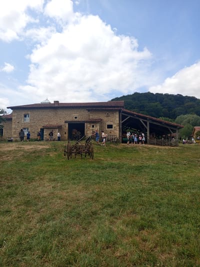 The image captures an outdoor scene at a historical reenactment or heritage site in Azannes-et-Soumazannes, France, on a partly cloudy day. A stone building with a red-tiled roof dominates the background, featuring an open-sided covered area where several people, including children, are gathered. Two horses are visible within this covered area, behind a wooden fence. In the foreground, a group of individuals, seemingly visitors, are observing the site and interacting. Various antique farming implements are displayed, including a weathered wooden and metal plow in the middle ground and other machinery near the building. The environment consists of a grassy slope leading up to the structures, with a treed hillside forming the backdrop under a sky with scattered white clouds.