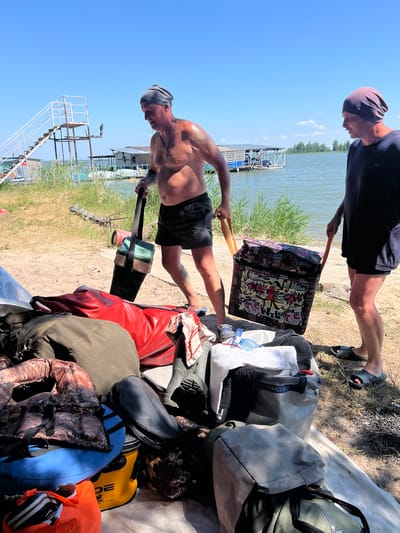 Two men are on a sandy shore next to a body of water on a sunny day. The man on the left, bare-chested and wearing a bandana and black shorts, carries a dark green and beige fishing tackle box with a black strap. The man on the right, wearing a dark blue shirt and black shorts, carries a large, colorful, patterned tote bag with orange handles. They appear to be moving luggage, as a pile of bags and items is located near them on the ground, including red and olive green duffel bags, a camouflage vest, a blue inflatable bag, a yellow bucket, and black boots. In the background, there is a wooden structure with stairs leading into the water, a floating platform with a building, and vegetation along the shoreline. The water is calm, and the sky is clear blue with a few scattered clouds. The location is a beach near Borovoye, Russia.