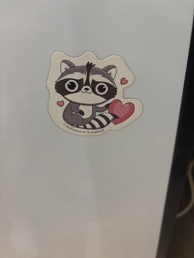 This media file shows a sticker of a cartoon raccoon holding a heart. The raccoon is depicted in shades of gray and black with large, round eyes and a striped tail. It is lying on its side, hugging a pink heart. The sticker has a white border and is placed on a light-colored, flat surface, possibly a desk or laptop. There are small pink hearts around the raccoon. To the right of the raccoon, there is text written in a circular arc. The text reads "с любовью от b.makeup". The overall image conveys a cute and affectionate theme. The sticker is likely a promotional item or a decorative decal. The location context provided, Княгинино, Russia, suggests the origin or intended distribution of this item.