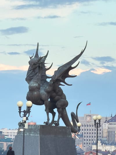 This image depicts a statue of a mythical creature, possibly a dragon or griffin, made of dark metal. The statue is positioned outdoors against a backdrop of a bright blue sky with scattered white and pinkish clouds. In the background, a cityscape is visible, featuring tall buildings with a distinct architectural style, suggesting an urban setting. A flag with red and white stripes is flying from a pole in front of one of the buildings. There are also streetlights with white globes, one of which is in the foreground, partially obscuring the statue. A person, seen from the back and mostly out of focus, is walking on the right side of the image. The lighting suggests it is daytime, and the clear sky indicates fair weather. The composition is vertical, and the image appears to be rotated, with the sky at the bottom and the ground at the top. No visible text is present in the image. The overall scene evokes a sense of grandiosity due to the imposing statue and the urban backdrop.