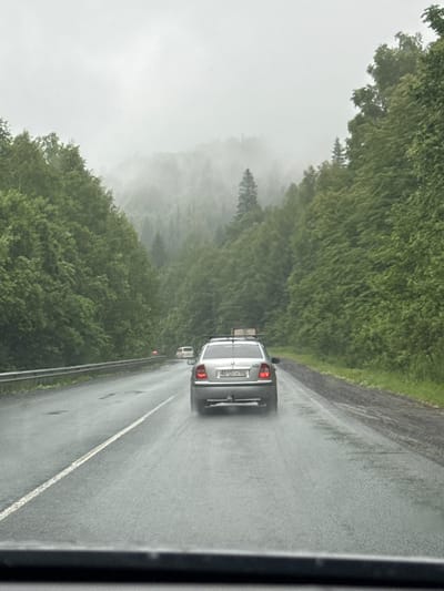 The image depicts a road scene in Brishbash, Russia, characterized by overcast skies and a light rain. A silver sedan is driving away from the camera on a wet, two-lane road bordered by dense green forest. The road surface appears to have some potholes and is covered in a thin layer of water. The forest on either side of the road is lush and extends up a misty, forested hillside. In the distance, other vehicles can be seen on the road. The overall atmosphere is subdued and damp due to the weather.