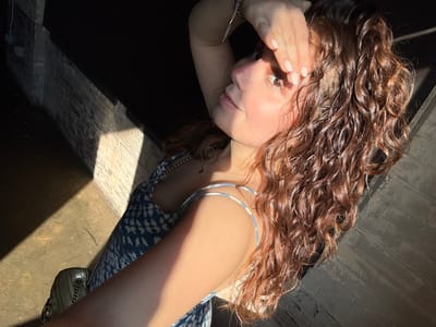 A woman reclines in dappled sunlight. Her curly, shoulder-length hair frames her face. She wears silver hoop earrings and a blue patterned dress. A purse with a chain strap rests on her shoulder. The sun highlights her face as she looks towards the camera, her hand near her cheek. A dark area is visible in the background, contrasting with sunlit walls.