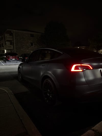 This is a nighttime, eye-level, medium shot of the rear passenger side of a dark gray Tesla Model Y parked on asphalt next to a curb. The car's brake lights are illuminated, emitting a bright red glow. In the background, to the left of the Tesla, a dark red SUV is parked in a lit parking spot. Further in the background, a brick apartment building with balconies and vine-covered walls is visible. Trees are present on the right side of the frame. The overall lighting suggests it is dark, with artificial lights from the apartment building and parking lot illuminating portions of the scene. There are no people visible in the image. The location is identified as Vernon Hills, United States of America.