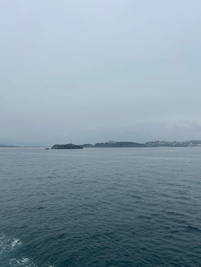 The image displays a scenic coastal view from a water-based perspective, likely a boat. The scene is divided vertically, with the left side dominated by a hazy, overcast sky and the right side by the calm, textured surface of the sea.
In the distance, along the horizon, a faint outline of a coastline is visible. This coastline appears to be a developed area, with buildings suggesting a town or city nestled amongst trees and hills. Further down the coast, a prominent rocky outcrop juts into the sea, topped with a small, white structure resembling a lighthouse or a monument. The water is a deep, muted blue-gray, with gentle ripples creating a textured surface. Near the bottom right corner, some white foam indicates the boat's wake.
The weather appears overcast and possibly foggy, contributing to the soft, diffused light and the hazy appearance of the distant land. The time of day is not distinctly clear due to the overcast conditions, but it seems to be daytime. The overall mood is tranquil and slightly somber due to the muted colors and lack of direct sunlight. No people or specific objects are clearly identifiable beyond the distant lighthouse-like structure. No text is visible in the image. The provided location context places this scene in Ribamontán al Mar, Spain.