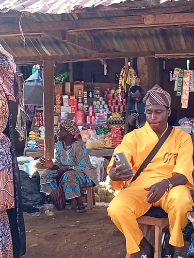 The image depicts an outdoor market stall in Wamba, Nigeria. In the foreground, a young man wearing a bright orange outfit and a striped head covering is seated on a wooden stool, looking at his smartphone. He has a dark strap across his shoulder and a watch on his left wrist. To his left, an older woman is also seated on a stool, dressed in a brown and blue patterned garment and a headscarf. She is looking to her right and appears to be speaking, with her hands gesturing. Behind them, another man in a dark shirt is visible, also looking at a smartphone. The stall itself is laden with various goods, including canned food items, bags of snacks, and other packaged products, arranged on shelves and tables. The roof of the stall is made of corrugated metal and wooden beams, with some fabric material visible. The ground is dirt, with some debris scattered around. The overall impression is a bustling local marketplace.
